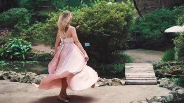 Pembe uzun elbiseli bir kadın dönüyor, dans ediyor, gülüyor ve gülümsüyor. Mutlu bir kadının olumlu duyguları. Arka planda yaz Batumi yeşil botanik bahçesi ile küçük bir gölet.