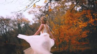 Beyaz elbiseli güzel sarışın poz veriyor. Ahşap sarısı yaprakların arka planı. Dans eden etek. Kumaş sinekleri el sallıyor. Olumlu duygular, çekici bir yüzün gülümsemesiyle mutlu olur. Sonbahar doğası