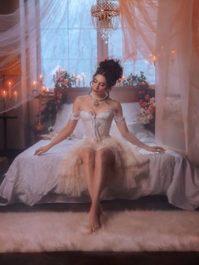 Seksi peri kız prenses manken yatakta poz veriyor güzel yüz dudaklar pembe makyaj yüksek saç stili koyu renk saçlar zarif asil bayan. Fantezi kadın kraliçesi, beyaz oda Viktorya dönemi tarzı elbise 