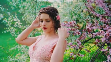 Mutlu gülümseyen kız pozları, çiçek açan arkaplan ağaçları, bahar mevsimi yeşil çimenler. Kadın gelin, zarif bir gelinlik, saç, yaratıcı tasarım saç stili, sanat diadeti.