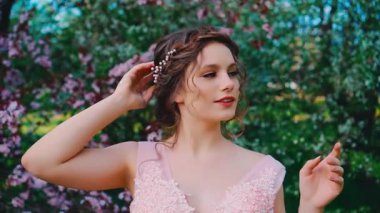 Mutlu gülümseyen kız pozları, çiçek açan arkaplan ağaçları, bahar mevsimi yeşil çimenler. Kadın gelin, zarif bir gelinlik, saç, yaratıcı tasarım saç stili, sanat diadeti..