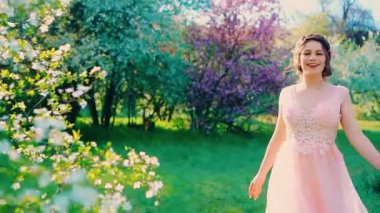 Güneşli parlak ışınlar parlıyor. Genç mutlu kadın bahar bahçesinde gülen yüzle yürüyor. Neşeli, neşeli, dans eden komik kız. Pembe elbise sallanıyor. Çiçek açan yeşil yapraklar