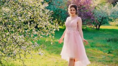 Güneşli parlak ışınlar parlıyor. Genç mutlu kadın bahar bahçesinde gülen yüzle yürüyor. Neşeli, neşeli, dans eden komik kız. Pembe elbise sallanıyor. Çiçek açan yeşil yapraklar