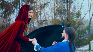 Tılsımlı kış ormanlarında, dallarda ve dallarda aşıklar buluştu. Yakışıklı ortaçağ esmer prensi, mavi eski moda kaşkort, el ele tutuşan güzel bir binici. Kırmızı kadife uzun lüks elbise, pelerin