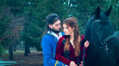 Romantik ortaçağ erkek prensi aşık kadın prensesi kucaklıyor. Seksi bayan siyah at hayvanı tutuyor. Fantezi klasik kırmızı elbise. Adam kral mavi takım. Yeşil kış doğa orman ağacı.