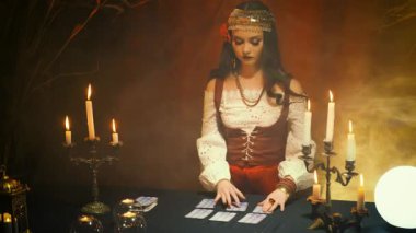 Karanlık gotik odadaki çingene cadı görüntüsünde güzel bir kız. Sanat elbisesi kırmızı kostüm. Falcı sihirli kadın tarot kartlarında geleceği okuyor kristal küre. Ayin mumlarının yanması, ruh çağırma dumanı.