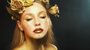 Kafasında gül çiçeklerinin yapay çelengi olan tanrıça figüründeki fantezi kadın portresi. Çelik gibi gri gözler yukarı bakar. Altın renkli boya damlaları güzel dudaklara damlıyor, kozmetik sıvısı dudak parlatıcısı.