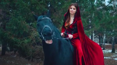 Fantezi kadın prenses siyah ata biner. Eski başlıklı bayan binici, kadife pelerinli kırmızı elbise. Yeşil ağaçlar orman kış doğası. Gülümseyen kız, evcil hayvanı sevgiyle okşar. 4k