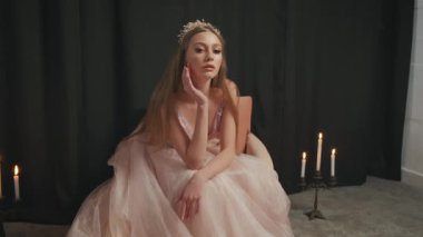 Altın tacın içindeki fantezi güzellik prensesi. Şamdanlar, mum ateşi yanıyor. Eski moda pembe şeftali elbiseli ortaçağ kadını. Kraliçe tahtta oturuyor. Moda modeli, siyah stüdyo.