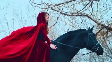 Ortaçağ kırmızı elbiseli prensesi siyah atlı ata biner. Eski başlıklı kız sürücü, kadife pelerinli tren rüzgarda uçuyor. Arka planda sonbahar ağaçları ormanları, mavi gökyüzü ilkbahar kış doğası