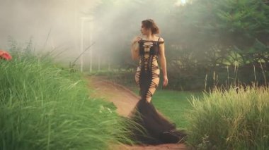 Genç seksi gotik kızıl saçlı, hayali bir kadının sisinde siluet, vücuda dokunuyor. Siyah, uzun, yaratıcı, baştan çıkarıcı elbise. Kara cadı ayakta duruyor. Arka plan uzun yeşil çimenler egzotik ağaçlar sisli dumanlar
