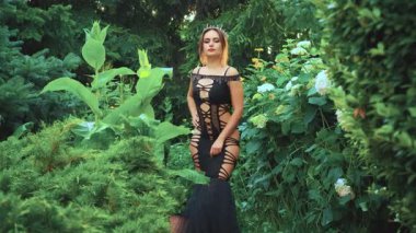 Fantezi genç kadın karanlık kraliçe seksi vücudu elleriyle okşuyor. Yetişkin kız prenses. Yaratıcı siyah vücutlu baştan çıkarıcı elbise kafasında gotik taç. Yaz bahçesi. Baharın yeşil yaprakları