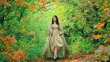 Kraliçe fantezi kadın yaz ormanlarındaki ağaçlar gibi mistik rüyalarda dolaşıyor. Yol üzerinde yeşil sarı turuncu yapraklar var. Ortaçağ prenses kız güzellik suratı. Kaliteli ipek elbise kabarık kollar uzun etek kenarı 4k