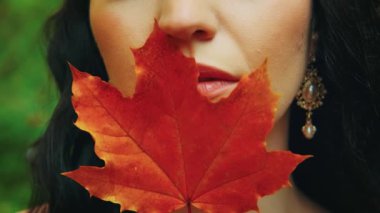 Ellerinde kırmızı sonbahar akçaağaç yaprağı tutan bir portre fantazisi kadın, Kız güzel gülen surat dudaklarını saklıyor. Yaz yeşili doğa. Altın küpeler klasik tarzda. Esmer, dalgalı saçlar. 4k