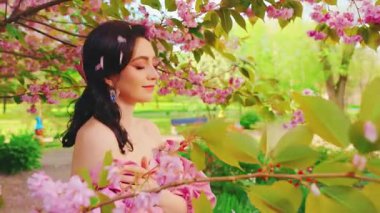 Bahar bahçesinde çiçek açan fantezi kız sakura yeşil ağacı. Güneş, mutlu kadın, güzel yüz, taze doğanın tadını çıkar. Rüzgarda uçan yapraklar. Pembe seksi elbise eski tarz. resim videosu