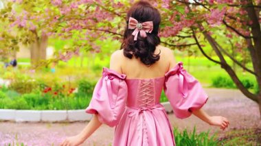Bahar bahçesindeki fantezi kraliçesi pembe çiçekler, parlak ağaçlar yaz parkı çimenleri. Uzun pembe elbiseli, ortaçağdan kalma eski moda kurdeleli bir kız saçının arkası manzaralı, doğanın tadını çıkarıyor. 4k