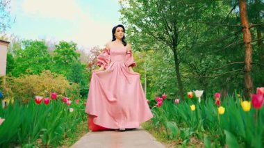 Fantezi kız prenses çiçek açan bahar bahçesinde yürüyor laleler yeşil çim yolu. Uzun pembe ipek elbiseli kadın kraliçe, kabarık kollu eski tarz. Aşk bekleyen romantik bir kadın. 4k