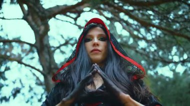 Cadılar Bayramı kostümü fantezi kadın portresi cadısı. Seksi vampir sihirbaz kız kameraya bakıyor, uzun saçlı başlıklı. Siyah kırmızı elbise, resim kıyafeti. Ağaç yeşili yapraklar orman, mavi gökyüzü