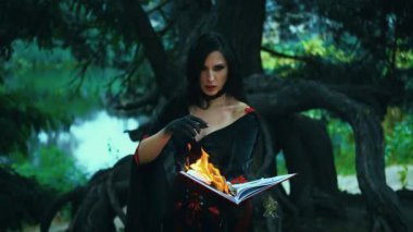 Fantezi kadın cadı eski yanan kitabı elinde tutuyor, parlak alev ateşi, kız gotik kız büyülü kırmızı elbise okuyor cadılar bayramı kostümü. Karanlık orman ağacı. Kötülüğe ışık, muhteşem yüz, gotik makyaj.