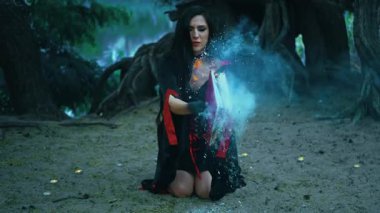 Fantezi seksi kadın cadı dizlerinin üzerine çöküp eski kitapları yakıyor, kapatıyor, kalın mavi duman çıkıyor, dişi gotik bayan siyah elbise cadılar bayramı cadılar bayramı kostümü. Gece ormanı ağacı