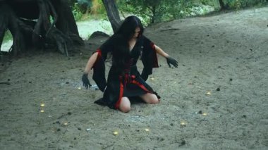 Fantezi portresi kadın cadı dansı ritüeli dansı. Seksi kız oturur sihirbaz elini gökyüzüne kaldırarak dua büyüsü yapar. Siyah kırmızı karnaval kostümü. Ağaç kökleri yeşil derin gotik orman nehri 4k