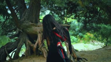 Cadılar Bayramı kızı Fantezi kadın cadı büyü ritüeli dansı. Seksi kötü kadın sihirbaz elini sıkar, uzun saçlar rüzgarda yavaş çekimde uçar. Siyah kırmızı elbise kostümü. Ağaç kökleri koyu yeşil orman 