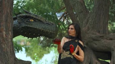 Portre sanatı fantezi kadın savaşçı kraliçe ejderhasını besliyor, düşmanının kalbini elinde tutuyor. Kara kraliçe, kafasını okşayarak hayvanın ağzına sevgiyle dokunur. orman yeşili ağaçlar. Siyah elbise 4K.