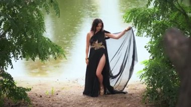 Uzun, zarif, ipek, siyah elbiseli fantezi gotik kadın yağmur altında nehir kıyısında yürüyor. Mistik kız prenses portresi. Yeşil ağaçlar yaz doğası. Vampir kadın ıslak siyah saçlar güzel yüz