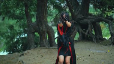 Fantezi portre kadın cadı ritüel dansı yapıyor. Seksi sihirbaz kız elini sıkar, uzun saç sineği ağır çekimde uçar. Siyah kırmızı karnaval kostümü. Ağaç kökleri yeşil orman suyu nehri