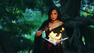 Fantezi kadın seksi cadı eski yanan kitabı elinde tutuyor, parlak alev ateşi, peri masalı gotik kız sihirli büyüyü okuyor. Cadılar bayramı için siyah kırmızı elbise. Karanlık orman ağacı. Kötülük yüzünde ışık