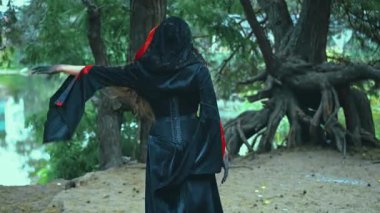 Gotik fantezi kadın büyücü yoğun bir çam ormanında yürüyor. Yeşil ağaçlar yaz doğası. Gizemli prenses. Klasik siyah kırmızı elbise, arka dikiz aynası, kafasında başlık. Ölülerin Vampir Kraliçesi