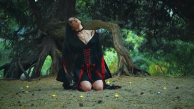 Fantezi portresi kadın cadı dansı ritüeli diz çökmüş oturuyor. Seksi kız sihirbaz başını sallıyor, uzun siyah saç sineği, düşük hareket. Siyah karnaval elbisesi. Ağaç gece ormanı yeşil doğa mumları yanıyor