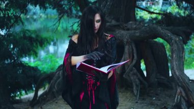Uzun saçlı, fantezi kadın seksi cadı eski kitap okuma büyüsü tutuyor. Kız peri masalı gotik vampiri. Cadılar bayramı için siyah kırmızı elbise. Karanlık orman ağacı. Eller siyah boyalı 4K