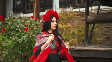 Evin yanında oturan mutlu kız portresi. Genç kadın Slav güzeli Ukraynalı siyah elbise kırmızı boncuklar, güzel uzun siyah saçlar. Bayan kendini çiçeğe sarar ılık eşarba soğuk sonbahar rüzgarına