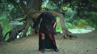 Fantezi portre kadın cadı ritüel dansı yapıyor. Seksi sihirbaz kız elini sıkar, uzun saç sineği ağır çekimde uçar. Siyah kırmızı karnaval kostümü. Ağaç kökleri yeşil orman suyu nehri