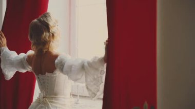 Genç kadın kraliçe perdeleri açar, pencereyi açar dışarı bakar yeni gün ışığının tadını çıkarır. Aşkı bekliyorum. Fantezi perisi bayan Mutlu kız sarı saç, beyaz klasik elbise eski tarz arka plan 4k.