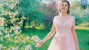 Güneş ışınları parlıyor. Genç, mutlu bir kadın bahar bahçesinde yürüyor, yüzünde gülümseme var. Neşeli, neşeli, dans eden komik kız. Pembe elbise sallanıyor. Çiçek açan yeşil yapraklar