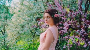 Genç güzel manken pozları, çiçek açan ağaçlar pembe beyaz çiçekler, bahar doğal yeşil çimenler. Kadın gelin, gelinlik, zarif elbise, saç, yaratıcı tasarım saç stili diyabeti.