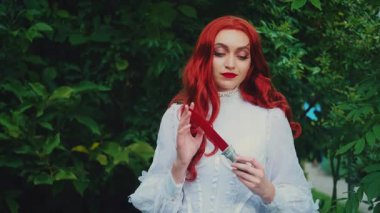 Fantezi portresi kızıl saçlı kadın kameraya bakıyor, cadılar bayramı makyajı kırmızı kanlı yüz izleri. Klasik beyaz elbise. Yaz yeşili ağaçlar. Gotik kız hançeri elinde tutuyor. Bıçakla oynuyor.
