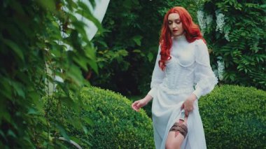 Fantezi kızıl saçlı tehlikeli kadın ortaçağ suikastçi kraliçesi. Deri kemerinde hançer olan. Beyaz, eski moda elbise. Kız prenses savaşçı. Yeşil yaz doğa ağaçları. Bıçağı tut..