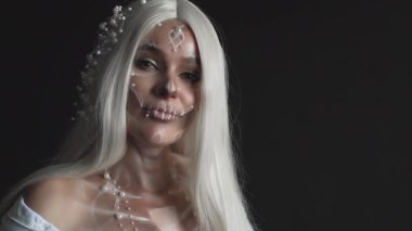 Beyaz saçlı, kolları kabarık ve omuzları açık beyaz elbiseli bir kadın. Yüzü inci takılı tatlı kafataslı bir kadın. Ölü gelin maskeli balosu için ölüm tanrıçası..