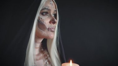 Gotik tarzda yüksek kaliteli video, okültizm ve fedakarlık, beyaz saçlı bir kadın siyah başlık takıyor ve elinde yanan bir mum tutuyor, yüzü boyalı kadın ölü iskelete benziyor..