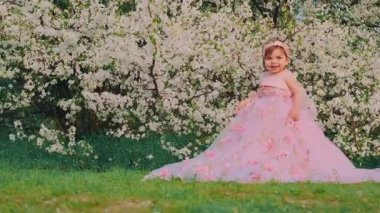 Küçük mutlu, güzel, neşeli, şen şakrak pembe elbiseli kız portresi. Gülen yüzlü çocuk prenses balo elbisesi tarzı. Arka planda bahar doğası çiçek açan beyaz ağaç yeşili çimenler