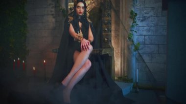 Portre fantezisi gotik kadın cadı tahtta oturuyor. Prenses kız seksi vampir kostümü, siyah pelerinli altın taç, uzun seksi bacaklar. Eski tarz oda tam sis dumanı, metal bronz demir sandalye