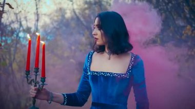 yaratıcı sanat video çekimi. Karanlık sonbahar ormanlarında portre fantezisi kraliçesi elinde yanan şamdanla kırmızı mumlar tutuyordu. Arka planda orman sonbahar ağaçları sis ve pembe duman kontrolörleriyle dolu.