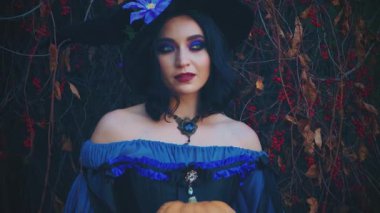 Cadılar Bayramı kadın cadı kostümlü mutlu yüzlü kadın turuncu balkabağı tutuyor. Gotik kız yaratıcı makyaj klasik mor elbise siyah külah şapka. Sonbahar doğası kırmızı ağaç yaprakları