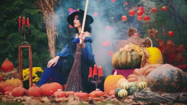 Ortaçağ 'ın güzel fantezi kadını oturur, elinde mistik sihirli süpürge tutar. Geleneksel balkabağı dekoru Cadılar Bayramı tatili. Mavi, klasik bir elbise, siyah bir şapka, çizgili çoraplar. kırmızı mumlar yanıyor, duman akıyor.