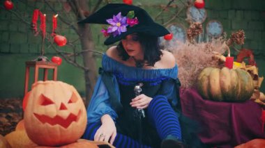 Modern, muhteşem, güzel, fantezi kadın bıçak tutuyor. Cadı güzel gülücüklerden, balkabağından, Cadılar Bayramı dekorundan hoşlanır. Mavi yaratıcı elbise, siyah külah şapka, sanat çiçekleri. Çıplak sonbahar dalları elma meyveleri 