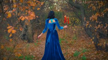 Güzellik sonbahar ormanı çıplak siyah ağaç gövdeleri. Gotik, zarif, fantezi kraliçesi. Kız vampir prenses elinde şamdanla, elinde yanan kırmızı mumlarla geri döndü. Uzun, mavi, eski bir elbise. Arka plan.