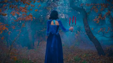Portre fantezisi korkmuş kadın kraliçe. Sonbahar karanlık ormanlarında gezen vampir kadın. Gotik, seksi, kötü kız, arkadan görünüşü, el ele tutuşan mumlar, yanan mumlar. Akan mavi sis sis bombaları. koyu ağaç